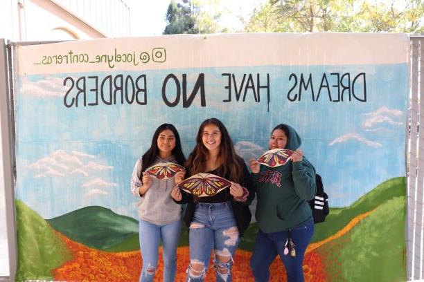 Three students holidng paper butterflies in front of the DREAMS HAVE NO BORDERS mural